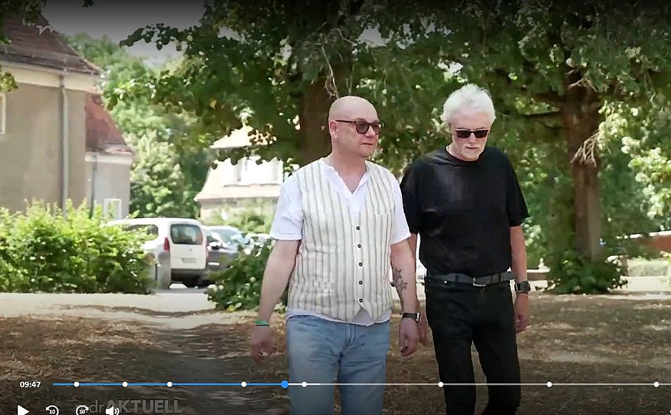 Zwei Männer spazieren nebeneinander in einem parkähnlichen Bereich; einer trägt eine gestreifte Weste, der andere ein dunkles Shirt und Sonnenbrille.