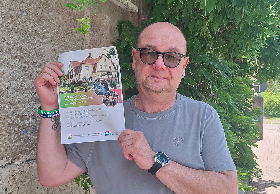 Mann mit Sonnenbrille hält einen Flyer vor einer Mauer mit Efeu, trägt graues T‑Shirt, Armbänder und Uhr.